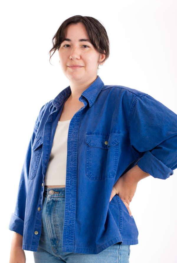 A person with short dark hair stands confidently with one hand on their hip. They are wearing a white tank top, a blue denim button-up shirt, and blue jeans. The background is white, creating a clean and simple backdrop.