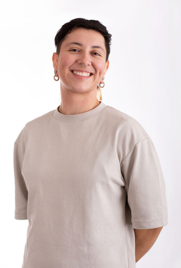 A person with short dark hair and hoop earrings smiles while standing against a plain white background. They are wearing a light-colored, short sleeve shirt and have their hands behind their back.