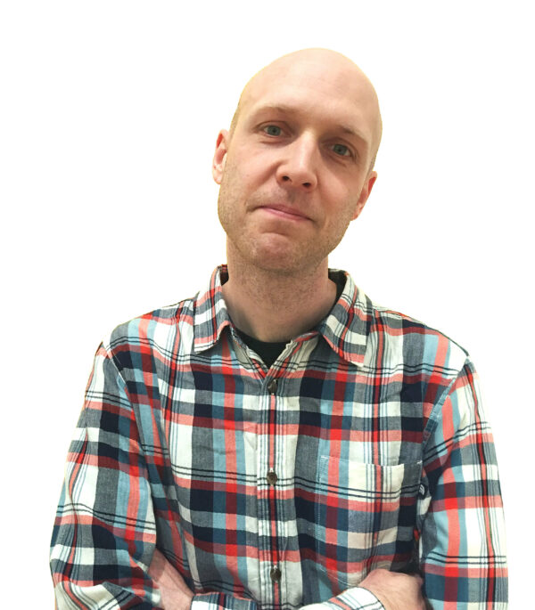 A person with a shaved head and a short beard is wearing a plaid shirt with red, blue, and white stripes. Their arms are crossed, and they are looking directly at the camera with a neutral expression. The background is plain white.