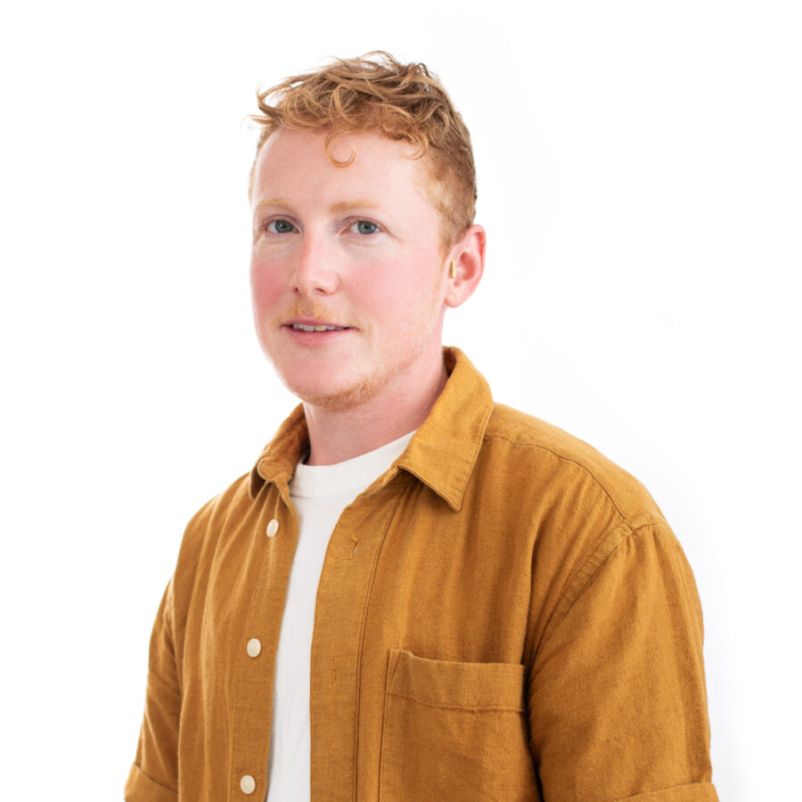 A person with short, curly red hair and light complexion is seated against a white background. They are wearing a mustard-colored button-up shirt over a white T-shirt and have visible tattoos on their arms. One hand is gesturing in front of them.