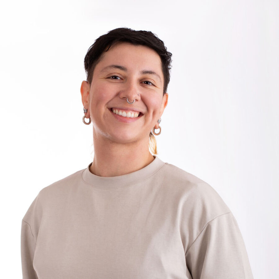 A person with short dark hair and hoop earrings smiles while standing against a plain white background. They are wearing a light-colored, short sleeve shirt and have their hands behind their back.