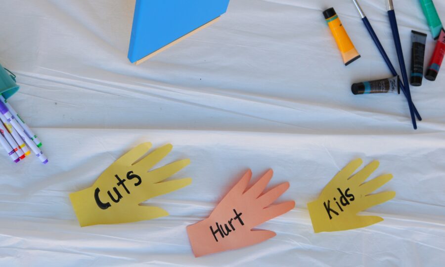 A table with art supplies, including paintbrushes, paint tubes, and markers, has three cut-out paper hands lying on it. The paper hands are yellow, orange, and pink, with the words "Cuts Hurt Kids" written on them.