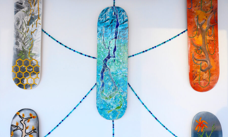 A collection of five vertical skateboards displayed on a wall. Each skateboard is uniquely decorated: one with a tree and octagonal patterns, one with a plant design, one with a blue river scene, one with an abstract tree, and one with red flowers. Blue beads connect the boards.
