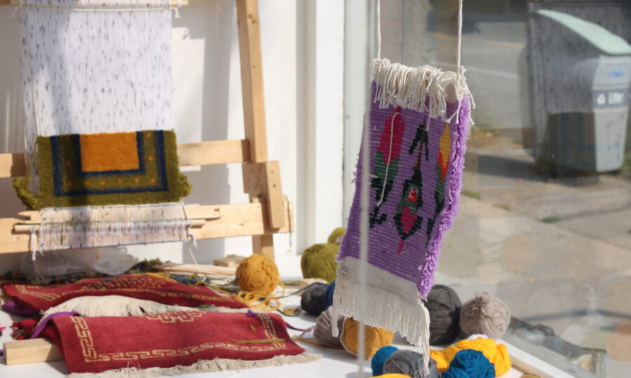 A workshop table displays handmade woven textiles, yarn balls in various colors, and some tools. A tapestry with a purple border and intricate patterns is hanging in the foreground, while other colorful weavings and a loom are visible in the background.