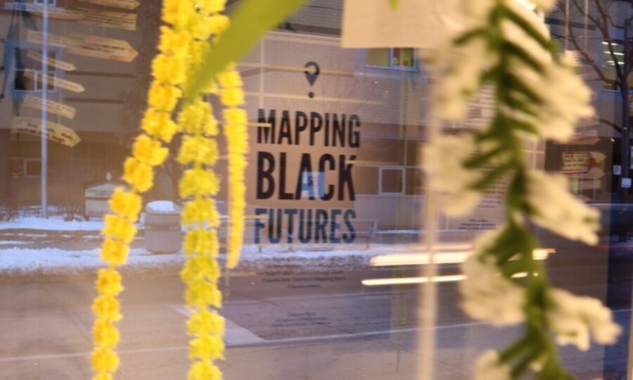 Yellow and white flower garlands are hanging in the foreground, partially obscuring a window with the text "Mapping Black Futures." A snowy street with buildings and trees can be seen through the window.