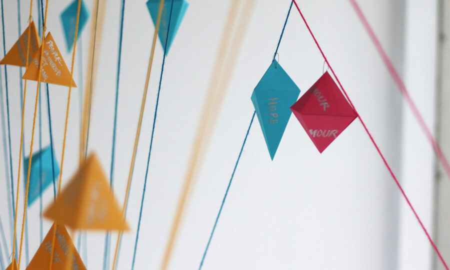 A close-up image of colorful paper shapes hanging on strings against a white background. The shapes include orange triangles, blue diamonds, and red diamonds, some with visible hand-written messages like "HOPE" and "YOUR" in white text.