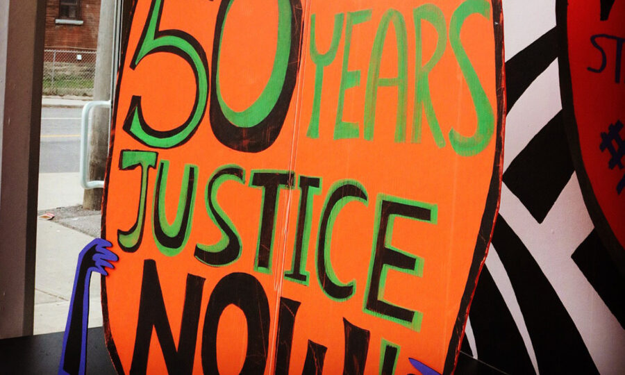 A bright orange sign with bold green and black letters reads "50 YEARS JUSTICE NOW!" Blue hand illustrations appear at the bottom of the sign. The background includes a street scene with a building and a sidewalk.