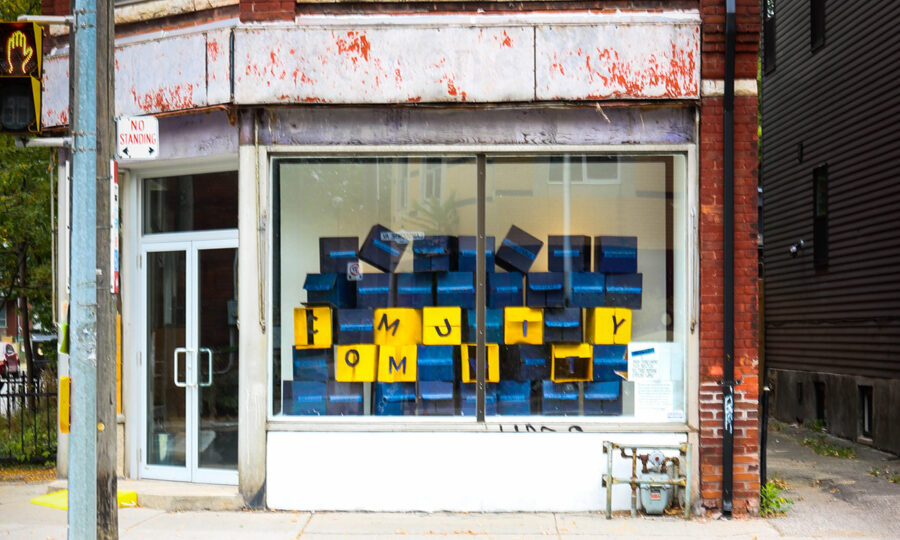 A storefront with large windows displays abstract blue paintings. Yellow letters near the bottom of the windows appear to spell out "FAMILY" among the art. The surrounding red brick building shows signs of weathering, and a "No Standing" sign is visible on the sidewalk.