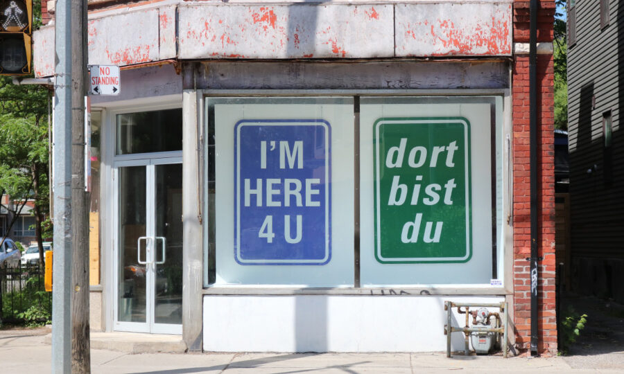 A street view of a building corner with windows displaying two large signs. The left sign is blue with white text saying "I'M HERE 4 U," and the right sign is green with white text saying "dort bist du." The building's exterior appears slightly worn.
