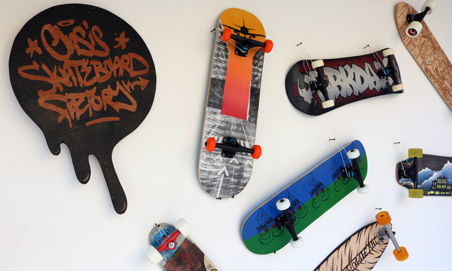 A white wall with various skateboards mounted on it. The skateboards have unique designs, including graffiti, landscapes, and abstract artwork. A sign shaped like a melting splash reads "Classic Skateboard Factory" in graffiti-style text.