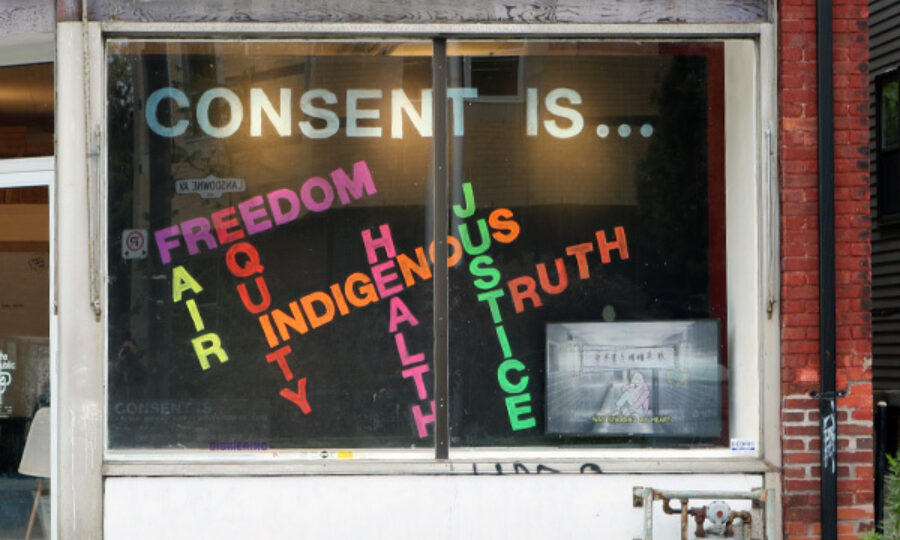 A shop window displays colorful letters forming the phrases "CONSENT IS..." followed by words such as "FREEDOM," "EQUITY," "INDIGENOUS," "HEALTH," "JUSTICE," "TRUTH." The angled letters are in shades of pink, orange, yellow, green, and red.