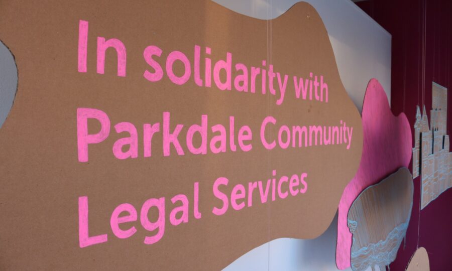 A cardboard sign with pink lettering reads "In solidarity with Parkdale Community Legal Services." The sign is displayed against a white wall with a pink abstract shape in the background.