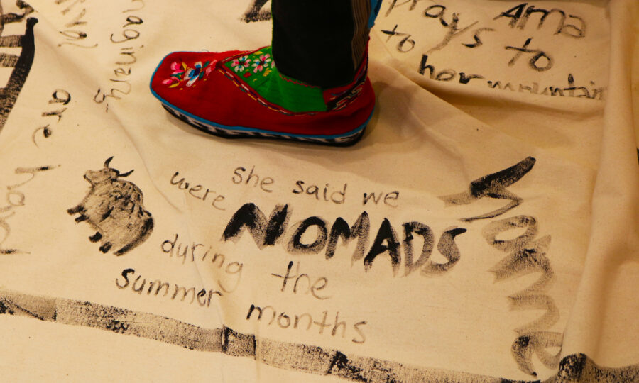 A close-up of colorful moccasin-clad feet standing on a cream-colored cloth. The cloth features handwritten text and drawings in black ink, including the words "she said we were NOMADS during the summer months" and an illustration of an animal.