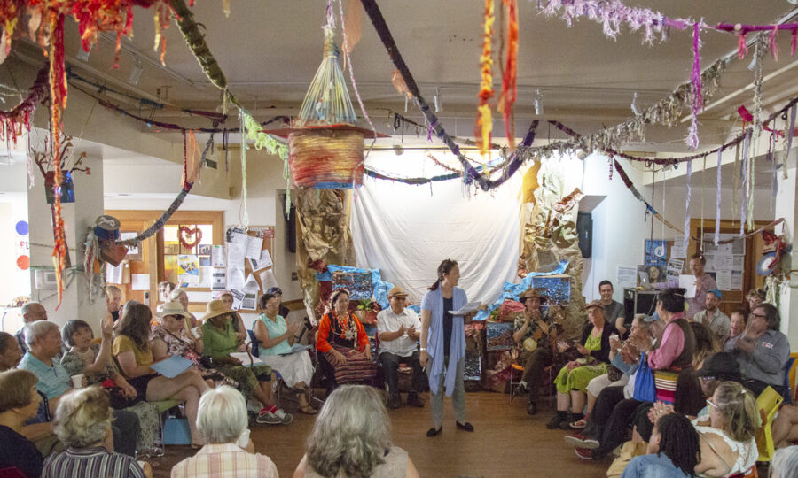 A large group of people sit in a circle in an eclectic room decorated with colorful yarn art hanging from the ceiling. A person stands in the center, speaking, while others listen attentively. The setting appears casual and artistic, with various decorations adorning the space.