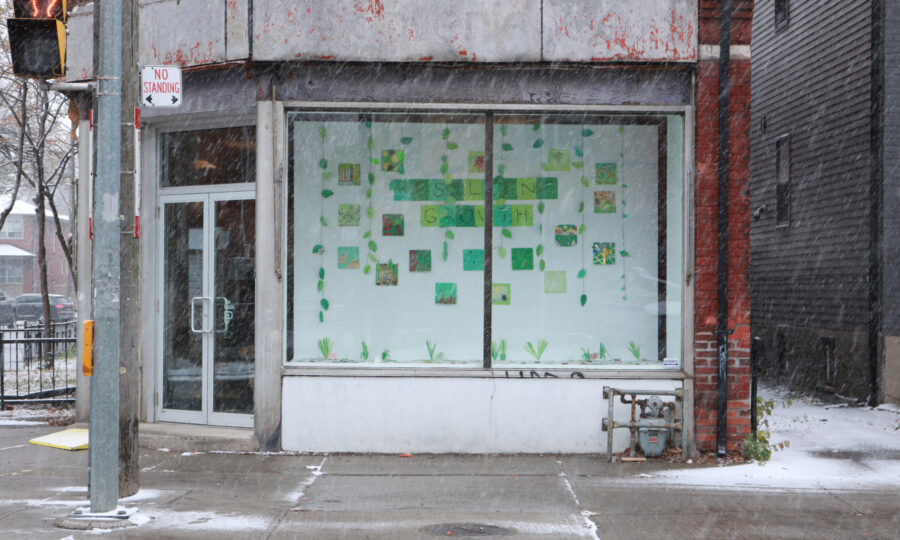 A street view of a corner building during snowfall. The building has a large window display featuring various green and yellow paper cutouts arranged on the glass. A "No Parking" sign is visible on a pole, and the ground is lightly dusted with snow.
