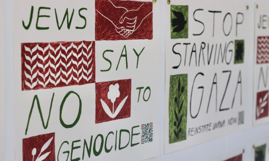 Three protest signs with artistic lettering and drawings. The prominent text reads: "JEWS SAY NO TO GENOCIDE" and "STOP STARVING GAZA." Background includes red and green designs with hand-drawn elements like hands shaking, a flower, and QR codes.