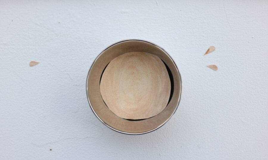 A round, empty metal container with a wooden base, viewed from above, is placed on a white surface. Two small teardrop-shaped marks are visible on the surface near the container, suggesting a minimalist or abstract design.