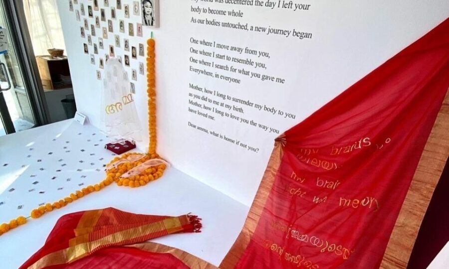 A red sari with gold details is displayed on a table near a white wall adorned with polaroids, yellow flowers, and various items. Text on the wall reads, "My world was decorated the day I left your body to become whole. Our bodies untouched, a new journey began.