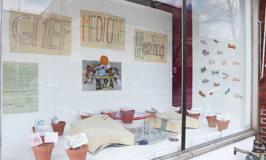 A storefront window display featuring potted plants with labels, photos, and colorful sticky notes. The background has a banner that reads "Grief, Medicine, Healing." The setup suggests themes of emotional healing and community engagement.