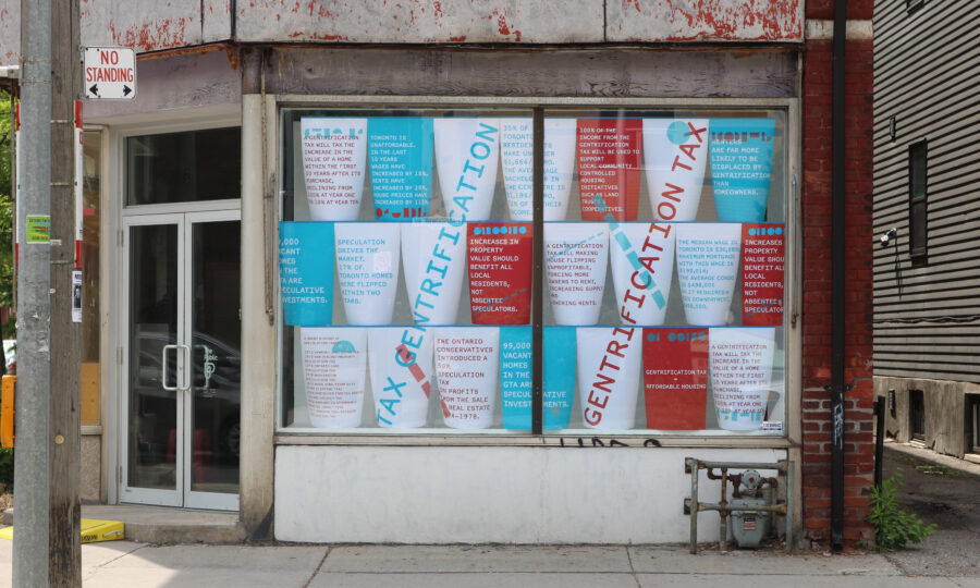 A storefront with a large window covered in colorful posters. The posters have text discussing issues like "gentrification" and "tax incentives." The exterior building is weathered with peeling paint. A "No Standing" sign is mounted on a nearby pole.