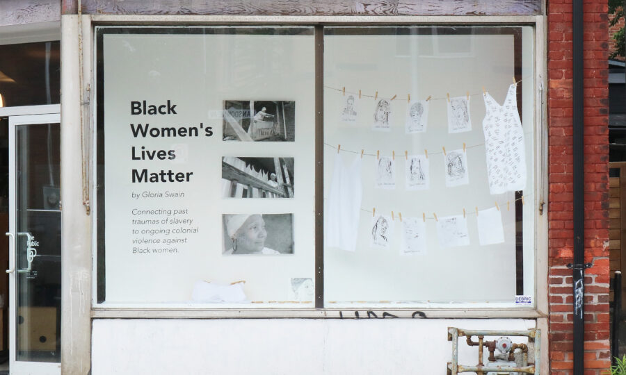 A storefront display features a white wall with the text "Black Women's Lives Matter" by Gloria Swain. Photos and artwork are pinned to strings across the window. The exterior shows an aged, red brick building and part of a sidewalk.